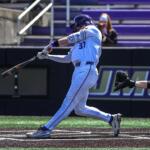 Baseball batter in a white uniform swinging at a pitch as the ball approaches, with the catcher in gray and black squatting behind him on the dirt field.