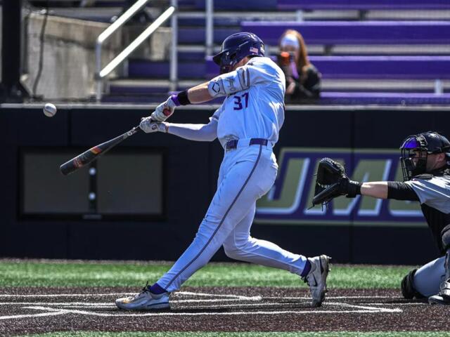 Baseball batter in a white uniform swinging at a pitch as the ball approaches, with the catcher in gray and black squatting behind him on the dirt field.