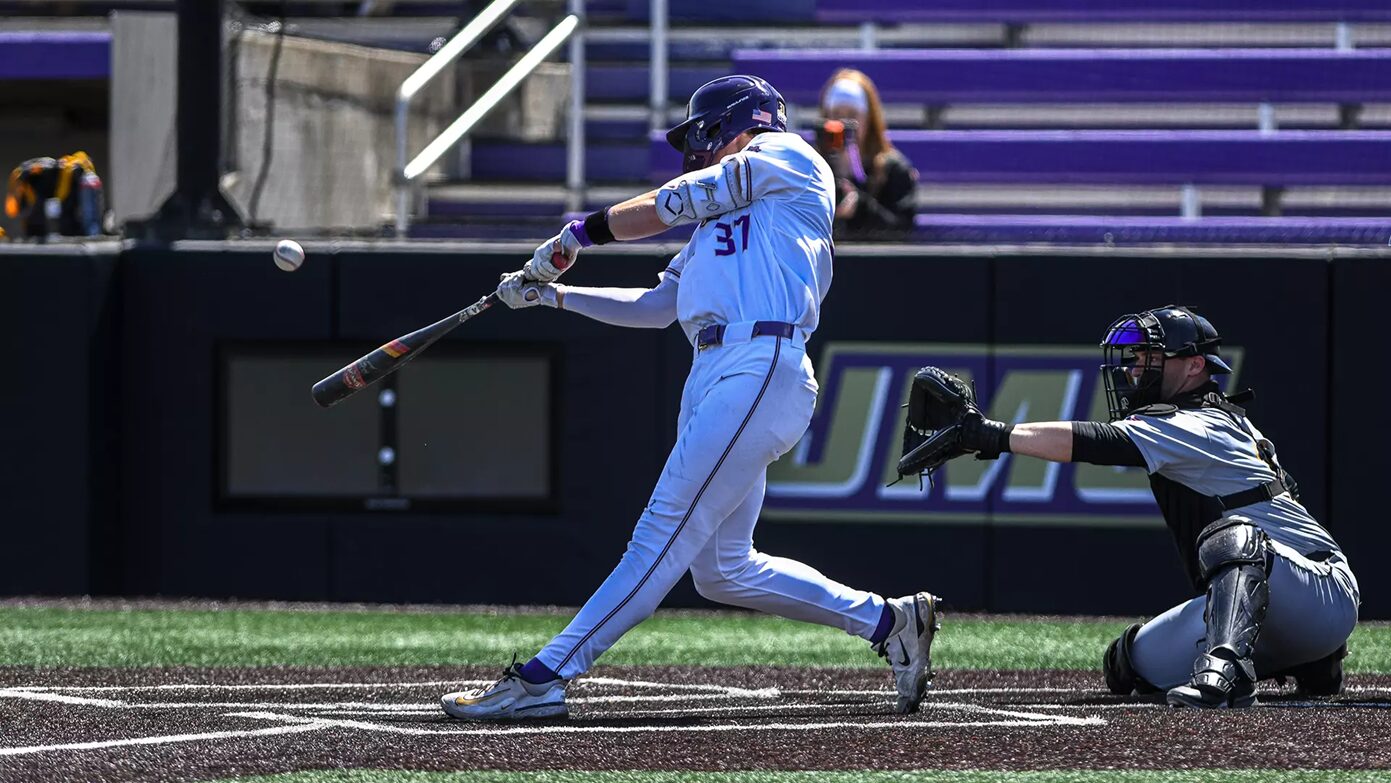 Baseball batter in a white uniform swinging at a pitch as the ball approaches, with the catcher in gray and black squatting behind him on the dirt field.