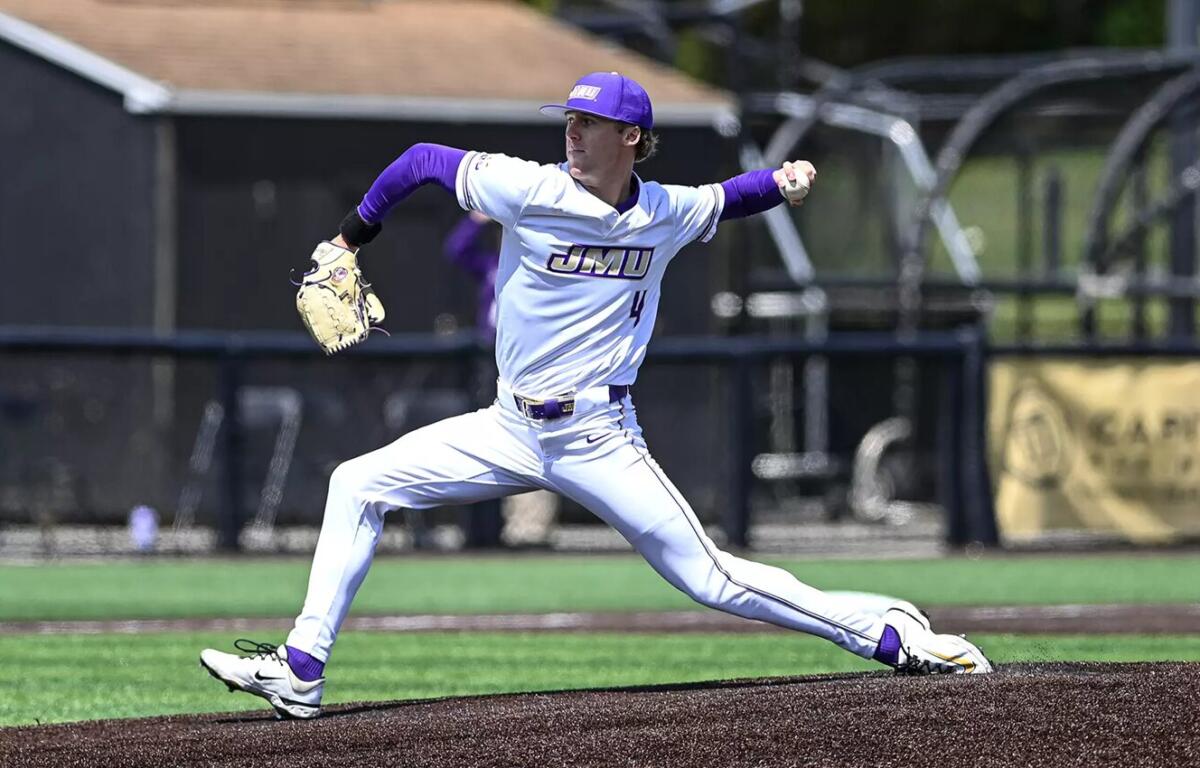 Male baseball pitcher in a white and purple JMU uniform delivering a pitch on the mound, left leg extended forward and glove raised.