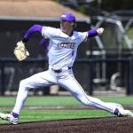 Male baseball pitcher in a white and purple JMU uniform delivering a pitch on the mound, left leg extended forward and glove raised.