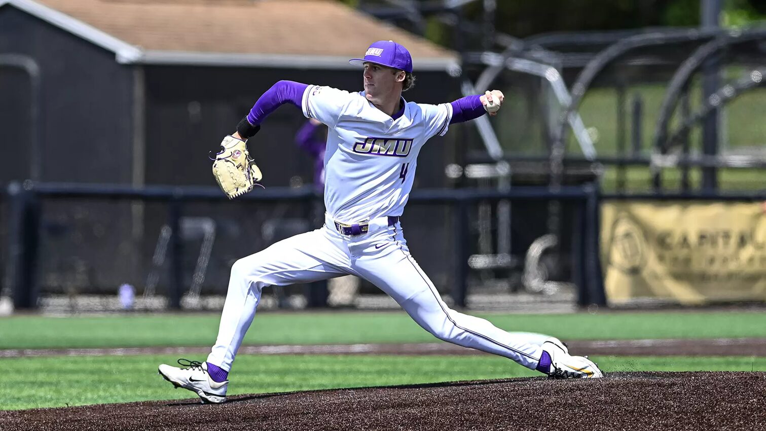 Male baseball pitcher in a white and purple JMU uniform delivering a pitch on the mound, left leg extended forward and glove raised.