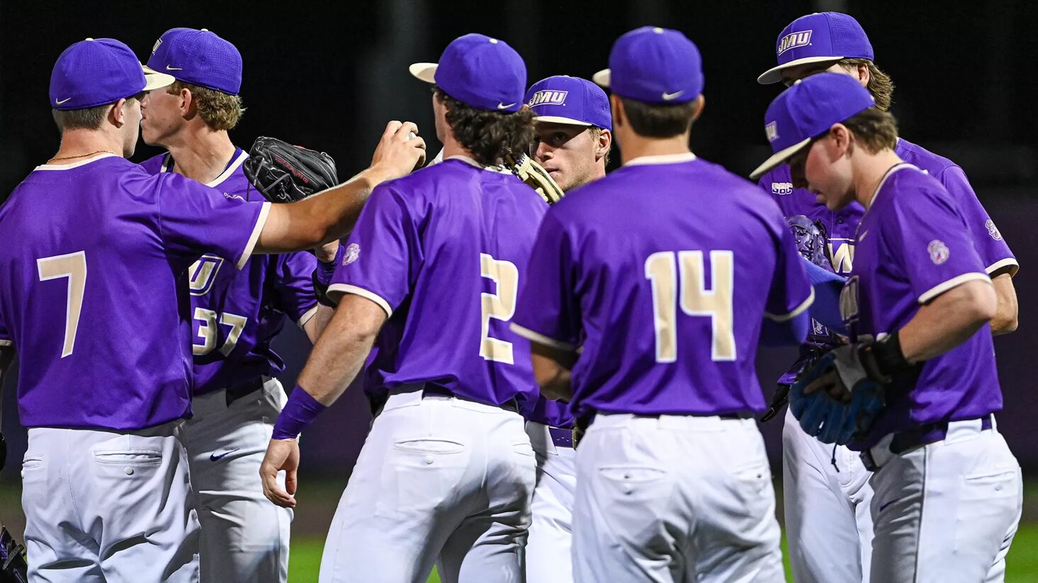 A group of baseball players in purple uniforms huddle on the field during a night game, backs to the camera with visible numbers 7, 2, and 14.