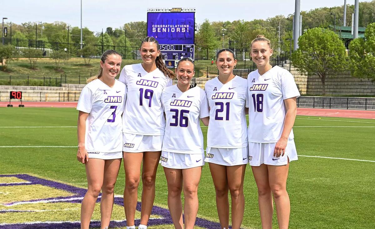 Five female soccer players in white JMU uniforms stand side by side on a sunny practice field, celebrating Senior Day with a purple scoreboard behind them.