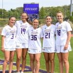 Five female soccer players in white JMU uniforms stand side by side on a sunny practice field, celebrating Senior Day with a purple scoreboard behind them.