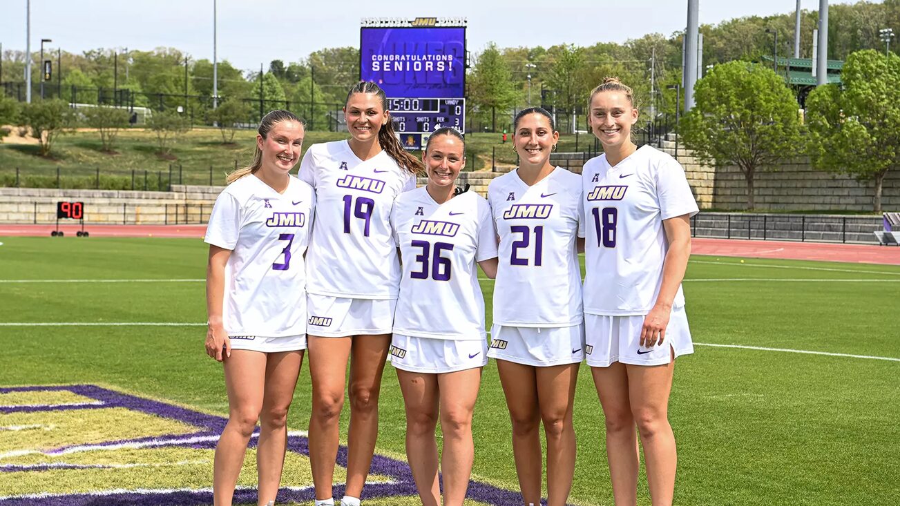 Five female soccer players in white JMU uniforms stand side by side on a sunny practice field, celebrating Senior Day with a purple scoreboard behind them.