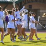 Group of female lacrosse players in white uniforms walk together on a sunlit field, high-fiving and carrying their sticks in celebration.