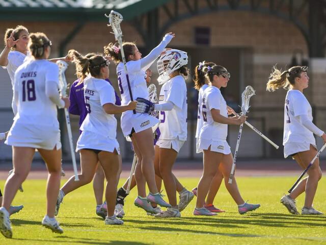Group of female lacrosse players in white uniforms walk together on a sunlit field, high-fiving and carrying their sticks in celebration.