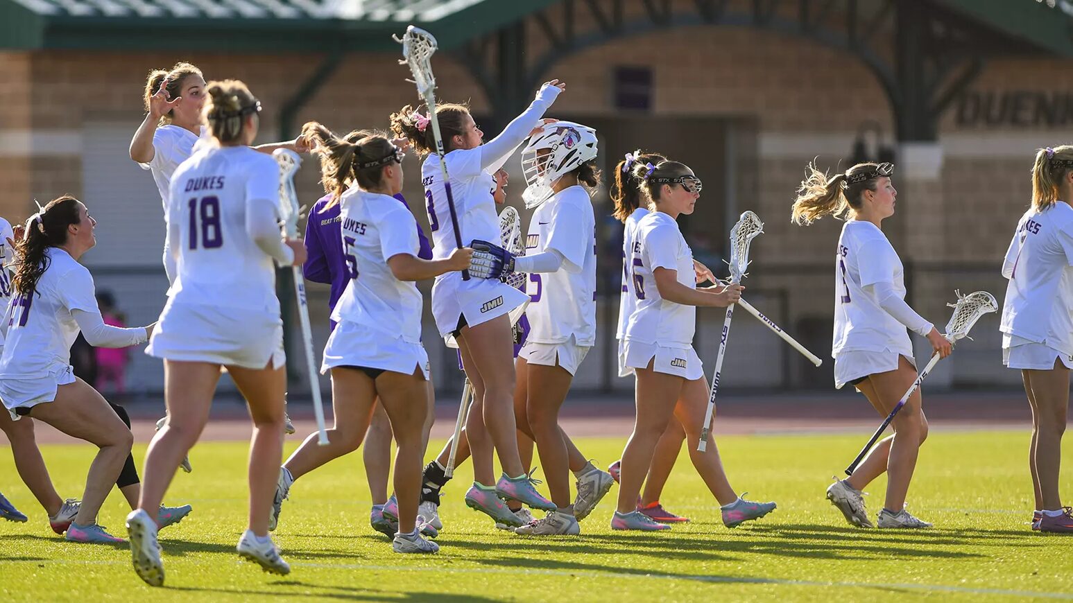 Group of female lacrosse players in white uniforms walk together on a sunlit field, high-fiving and carrying their sticks in celebration.