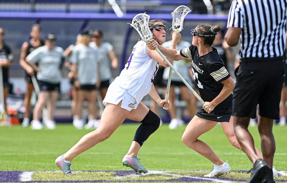 Two female lacrosse players collide for possession during a game on a turf field, one in white and one in black, near a referee in stripes.