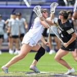 Two female lacrosse players collide for possession during a game on a turf field, one in white and one in black, near a referee in stripes.