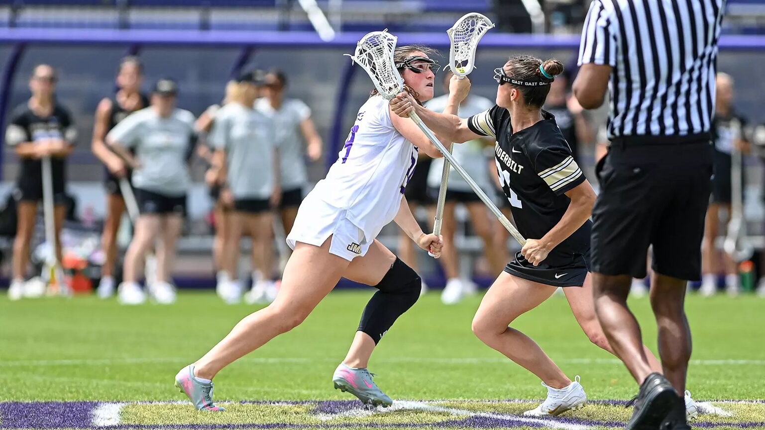 Two female lacrosse players collide for possession during a game on a turf field, one in white and one in black, near a referee in stripes.