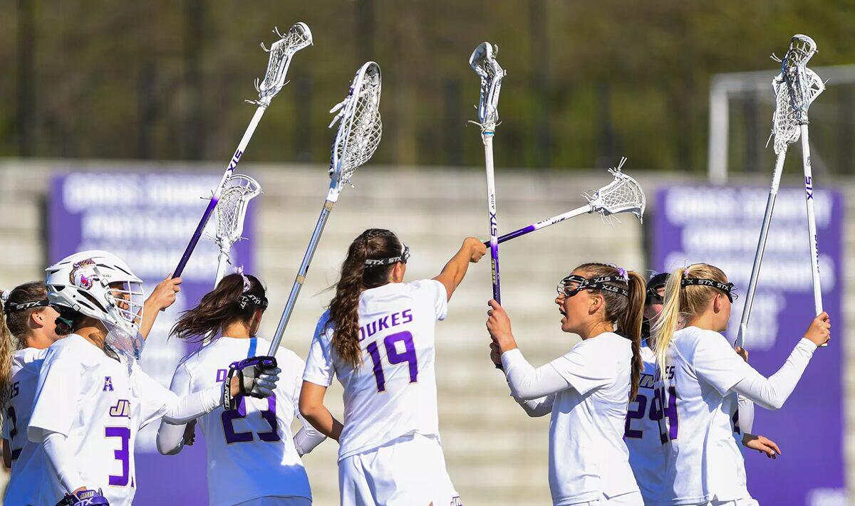 Lacrosse players in white uniforms celebrate on the field, raising their sticks in a high-five