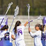 Lacrosse players in white uniforms celebrate on the field, raising their sticks in a high-five
