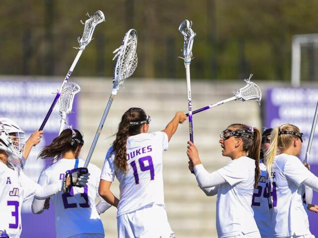 Lacrosse players in white uniforms celebrate on the field, raising their sticks in a high-five