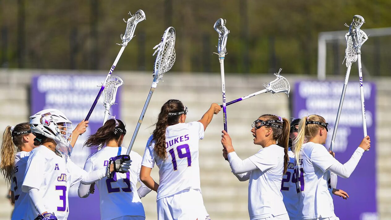 Lacrosse players in white uniforms celebrate on the field, raising their sticks in a high-five