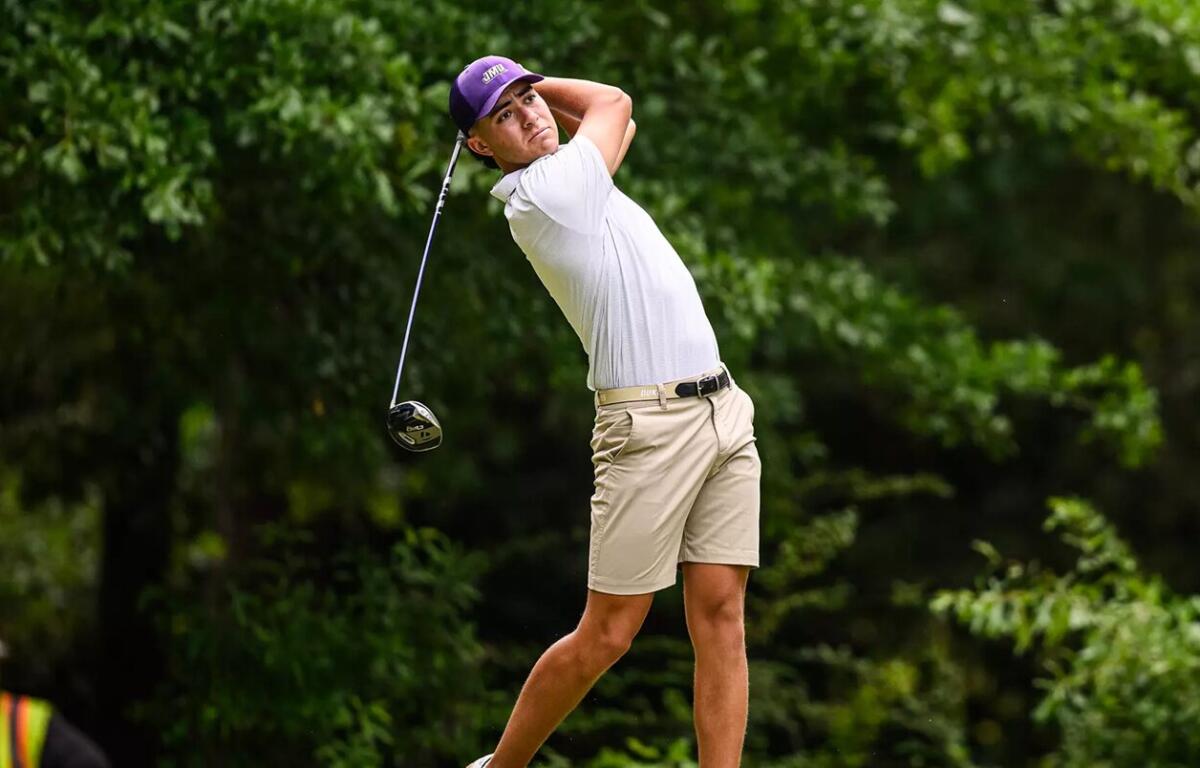 Male golfer mid-swing on a lush course, wearing a purple cap, white polo, and beige shorts with a driver raised behind him
