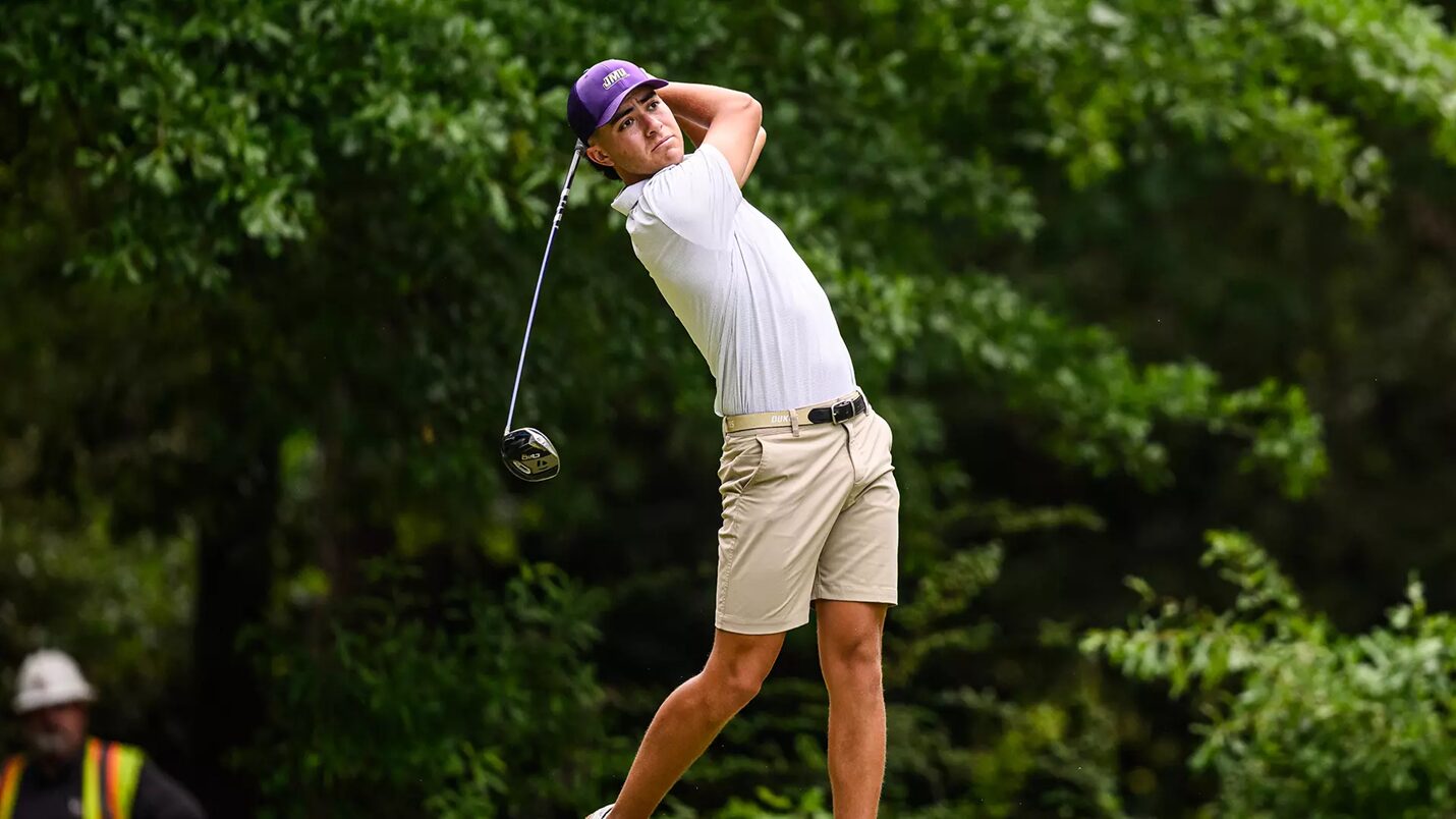 Male golfer mid-swing on a lush course, wearing a purple cap, white polo, and beige shorts with a driver raised behind him