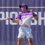 Tennis player in a purple cap and shirt hits a forehand during a championship match, ball near the racket on a blue court backdrop.