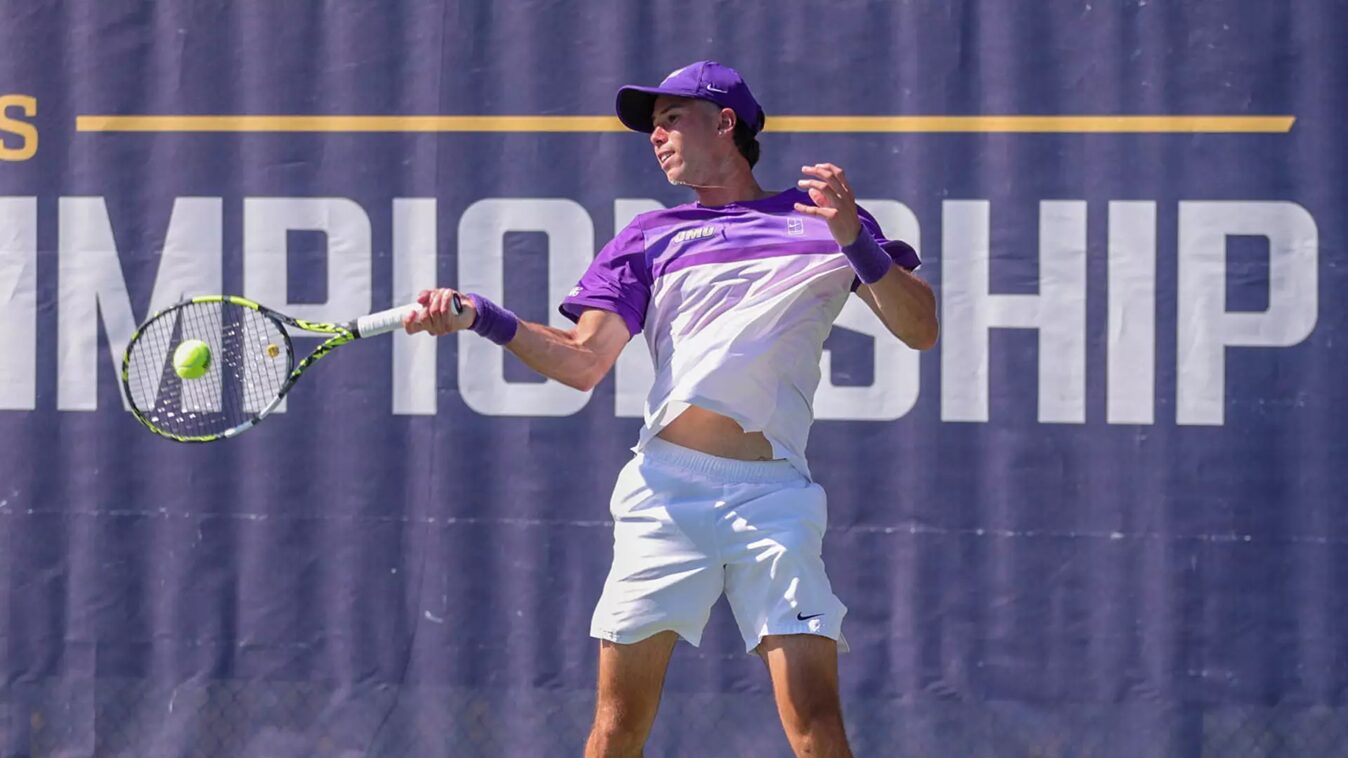 Tennis player in a purple cap and shirt hits a forehand during a championship match, ball near the racket on a blue court backdrop.