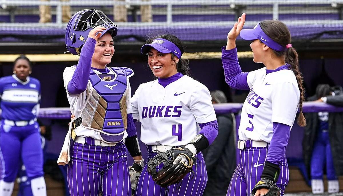 Duke softball players celebrate in the dugout, one wearing catcher gear and two teammates high-fiving and smiling in purple and white uniforms.