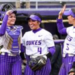 Duke softball players celebrate in the dugout, one wearing catcher gear and two teammates high-fiving and smiling in purple and white uniforms.