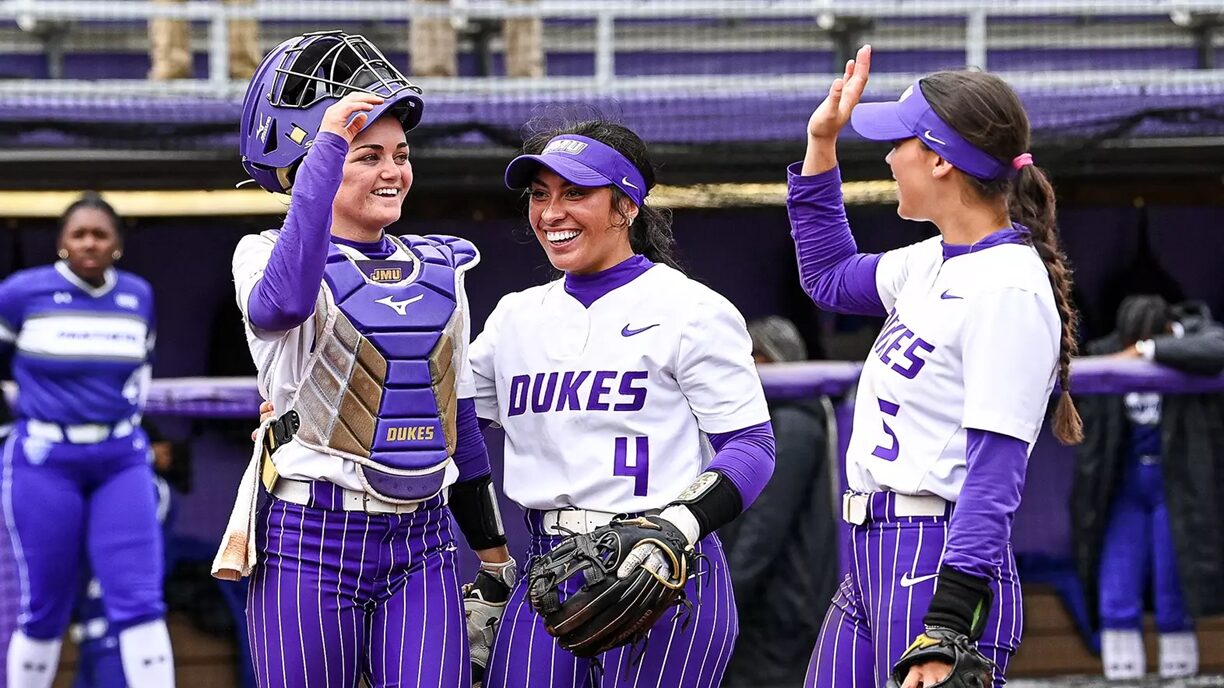 Duke softball players celebrate in the dugout, one wearing catcher gear and two teammates high-fiving and smiling in purple and white uniforms.