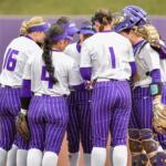 Girls' softball team huddling on the infield in purple pinstriped uniforms; visible numbers 16, 4, and 1; catcher wearing gear on the right.