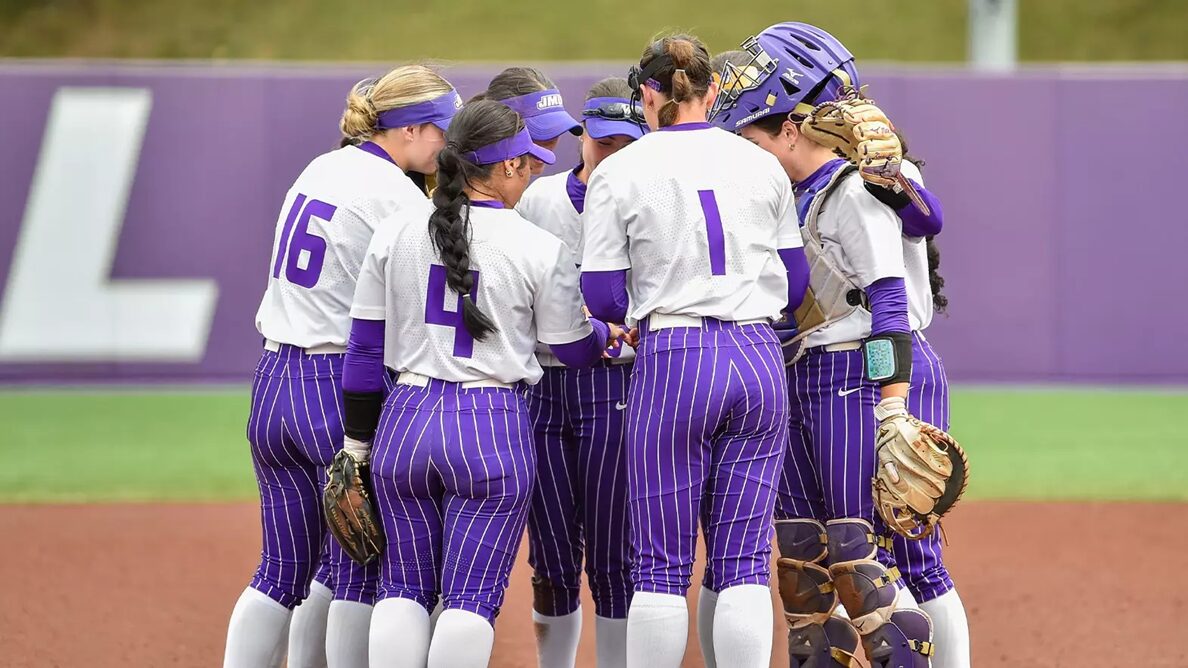 Girls' softball team huddling on the infield in purple pinstriped uniforms; visible numbers 16, 4, and 1; catcher wearing gear on the right.