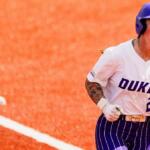 Dukes softball player sprinting on an orange infield, helmet on, fists clenched in celebration.