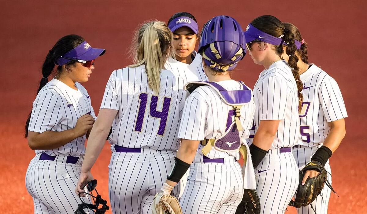 Softball players in white pinstriped uniforms with purple numbers huddle together on the field, one in catcher's gear visible in the back.