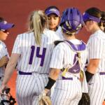 Softball players in white pinstriped uniforms with purple numbers huddle together on the field, one in catcher's gear visible in the back.