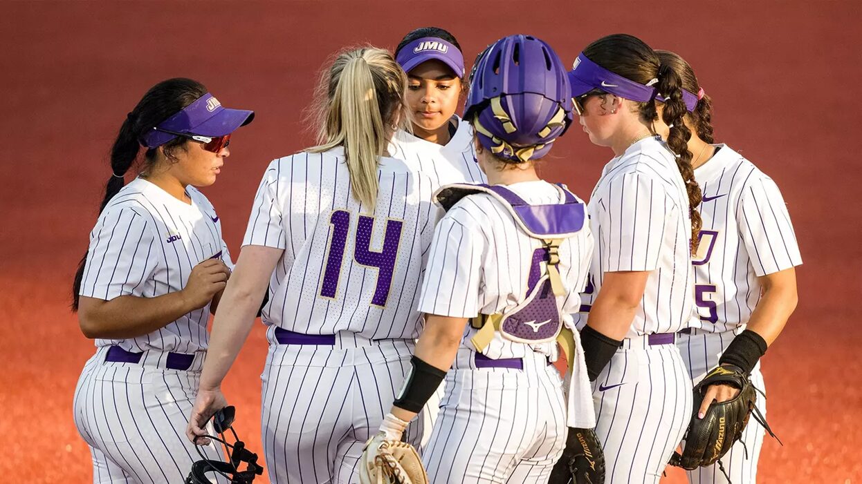 Softball players in white pinstriped uniforms with purple numbers huddle together on the field, one in catcher's gear visible in the back.