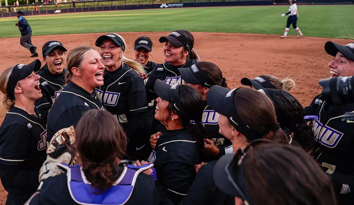 Softball team in black jerseys huddle in celebration on the infield dirt after a game.