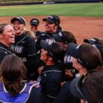 Softball team in black jerseys huddle in celebration on the infield dirt after a game.