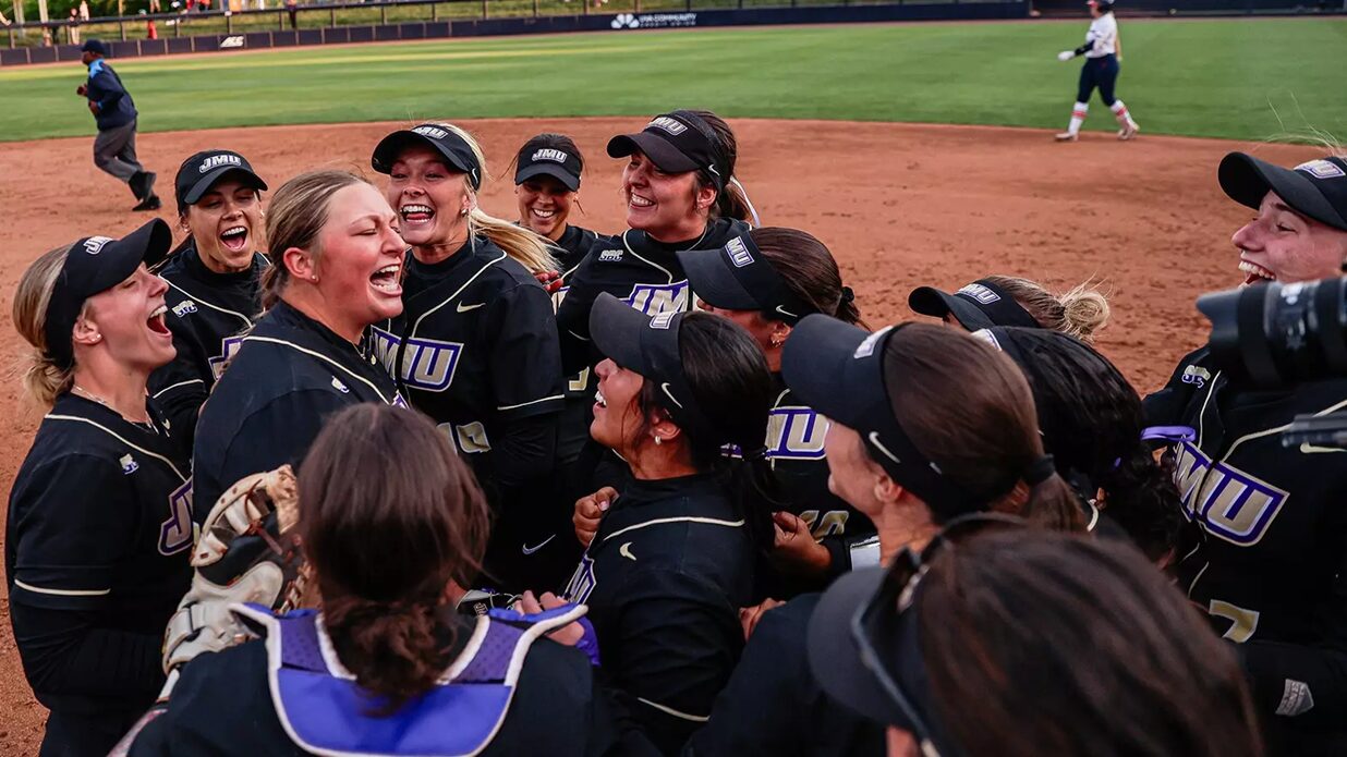 Softball team in black jerseys huddle in celebration on the infield dirt after a game.