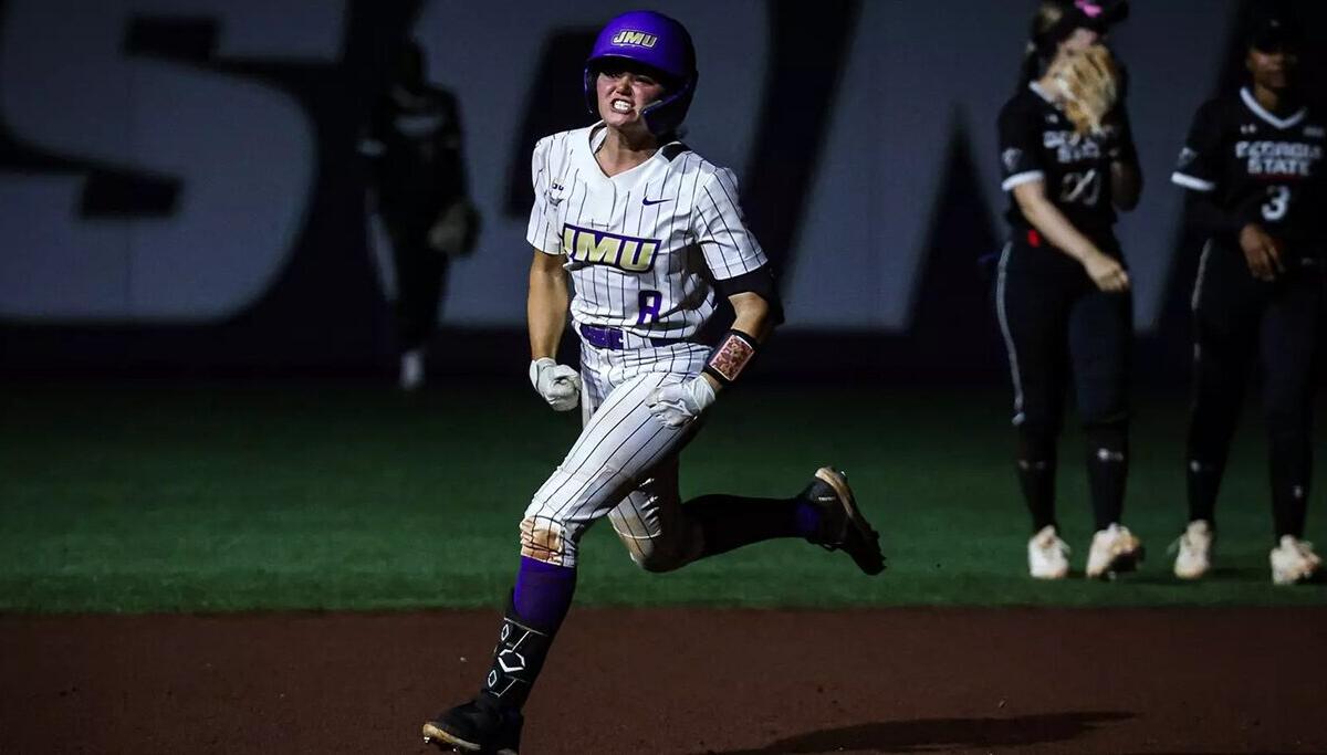 Female softball player in a white pinstriped uniform with purple accents runs on the infield at night, helmeted and focused.