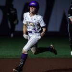 Female softball player in a white pinstriped uniform with purple accents runs on the infield at night, helmeted and focused.