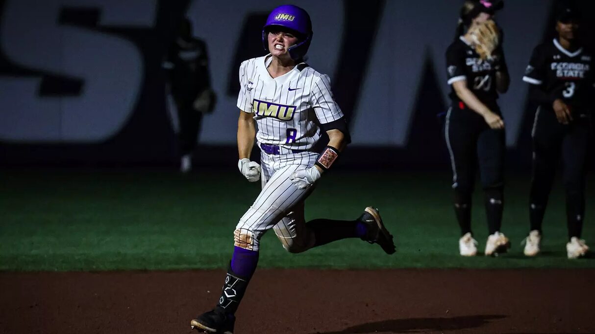 Female softball player in a white pinstriped uniform with purple accents runs on the infield at night, helmeted and focused.
