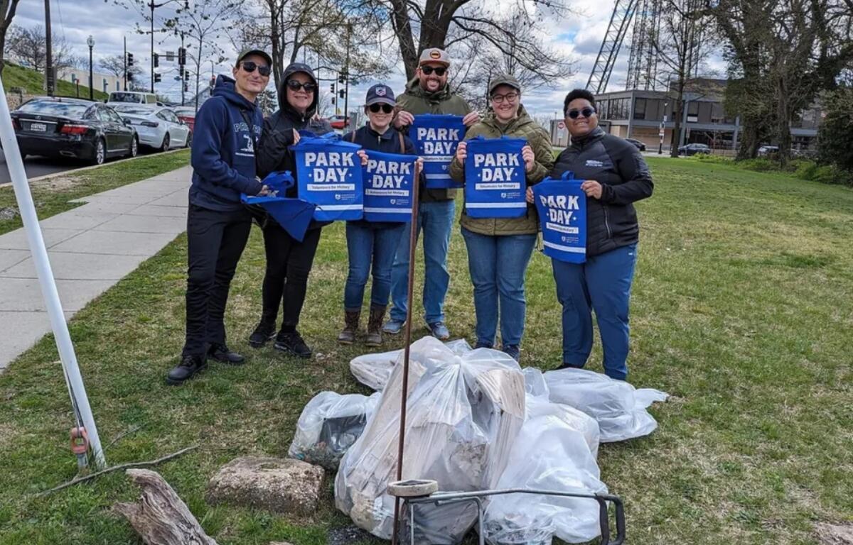 Six volunteers pose in a park, holding blue Park Day bags as they stand beside piled trash bags they've collected.