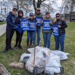 Six volunteers pose in a park, holding blue Park Day bags as they stand beside piled trash bags they've collected.