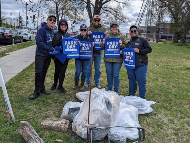 Six volunteers pose in a park, holding blue Park Day bags as they stand beside piled trash bags they've collected.