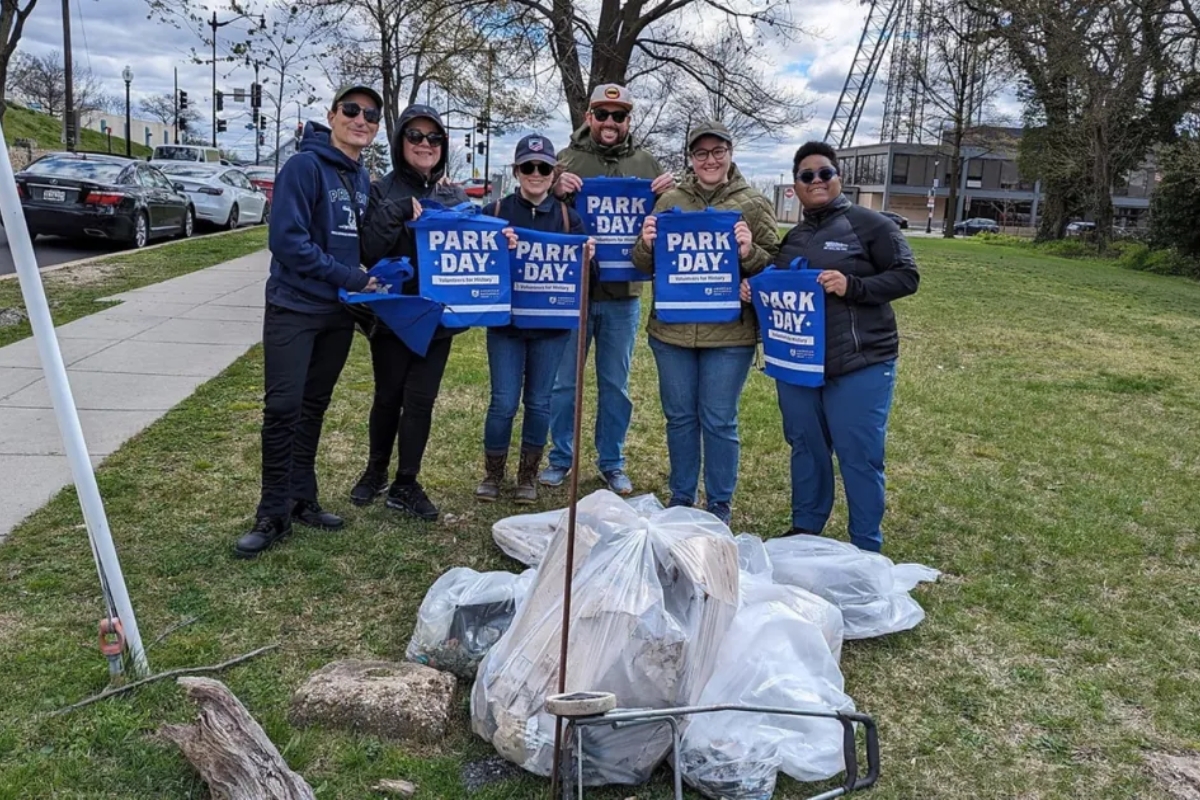 Six volunteers pose in a park, holding blue Park Day bags as they stand beside piled trash bags they've collected.