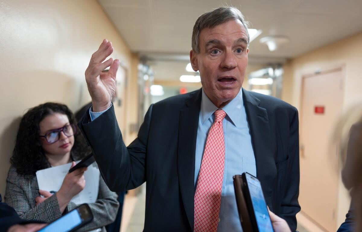 Man in a navy suit and red tie speaks to reporters in a hallway, raising his right hand as people hold phones and notebooks nearby.