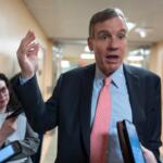Man in a navy suit and red tie speaks to reporters in a hallway, raising his right hand as people hold phones and notebooks nearby.