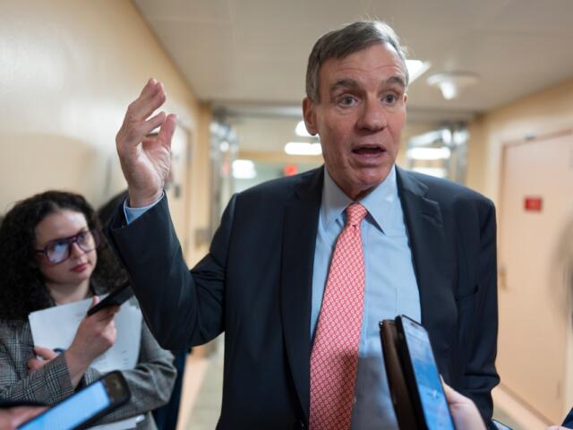 Man in a navy suit and red tie speaks to reporters in a hallway, raising his right hand as people hold phones and notebooks nearby.