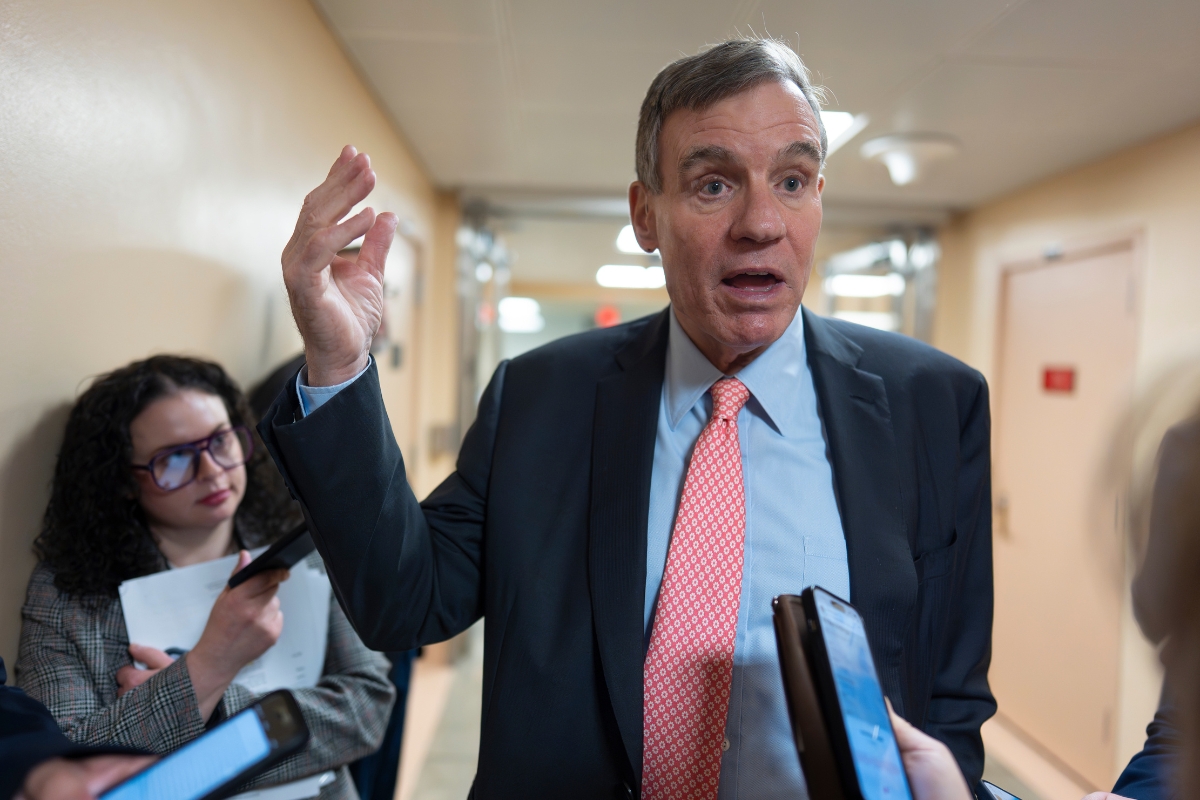 Man in a navy suit and red tie speaks to reporters in a hallway, raising his right hand as people hold phones and notebooks nearby.