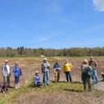 Group of hikers in a grassy field listening to a guide near a large boulder on a sunny day.