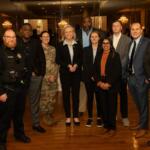 Group of ten professionals in business attire and security uniforms posing for a photo in a formal venue with chandeliers and mirrors in the background.