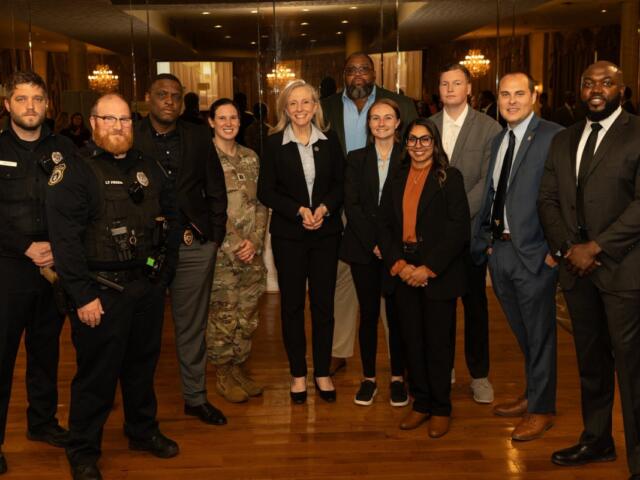 Group of ten professionals in business attire and security uniforms posing for a photo in a formal venue with chandeliers and mirrors in the background.